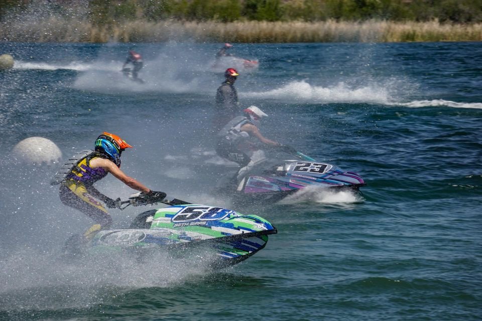 Jet ski racers speed across a blue lake, kicking up spray, with colorful personal watercraft and helmets visible.