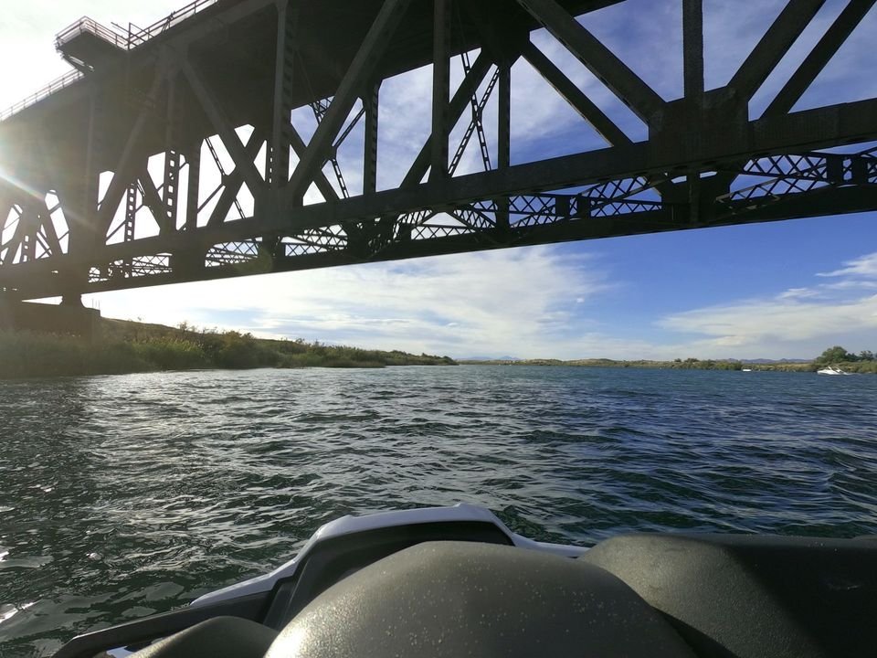 A view from a boat moving under a large metal bridge spanning a wide, blue river on a sunny day.