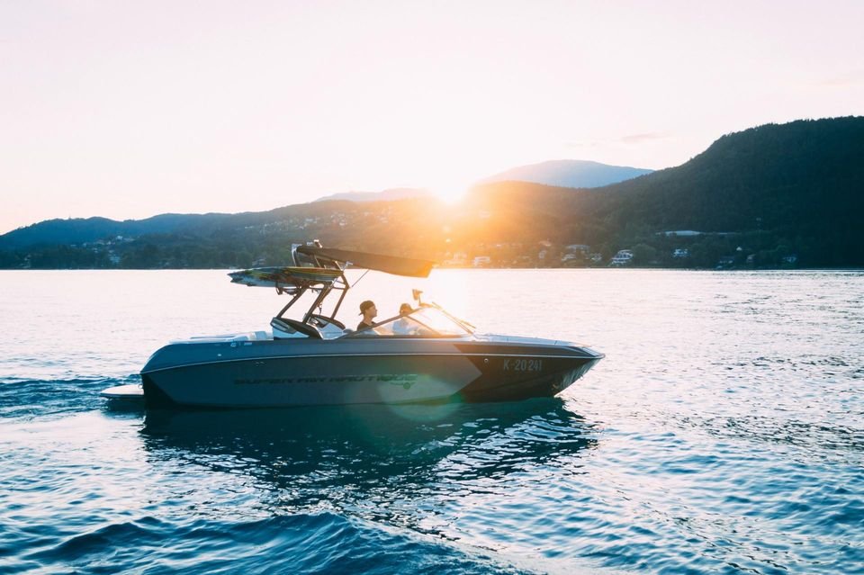 A motorboat carrying two people cruises across a calm lake toward a bright sunset behind distant mountains.
