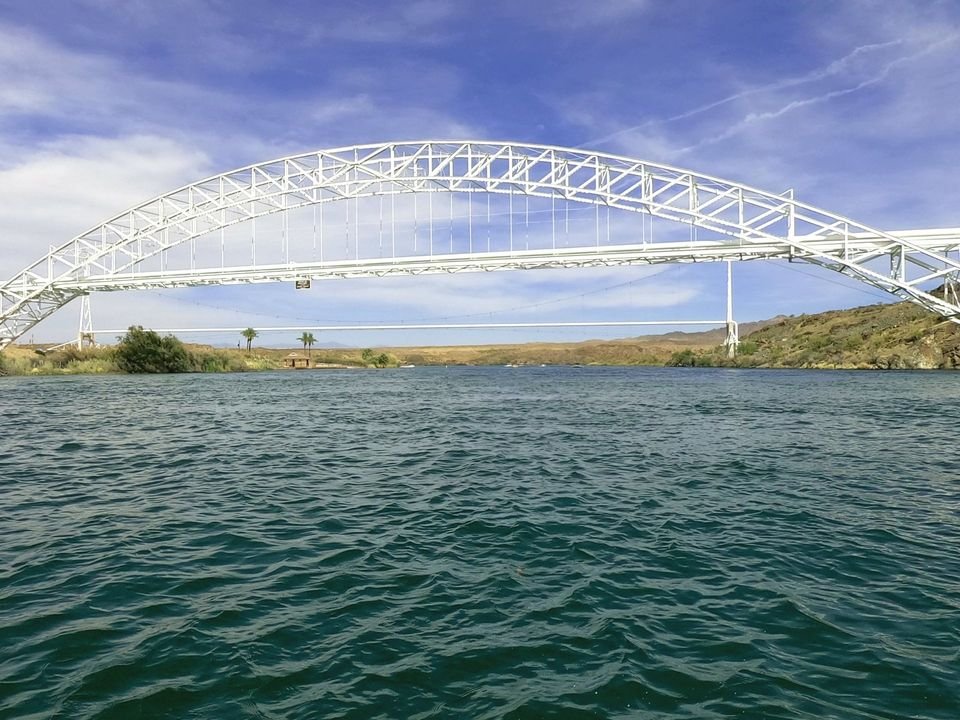A white steel arch bridge spans a wide, blue river under a bright, partly cloudy sky.
