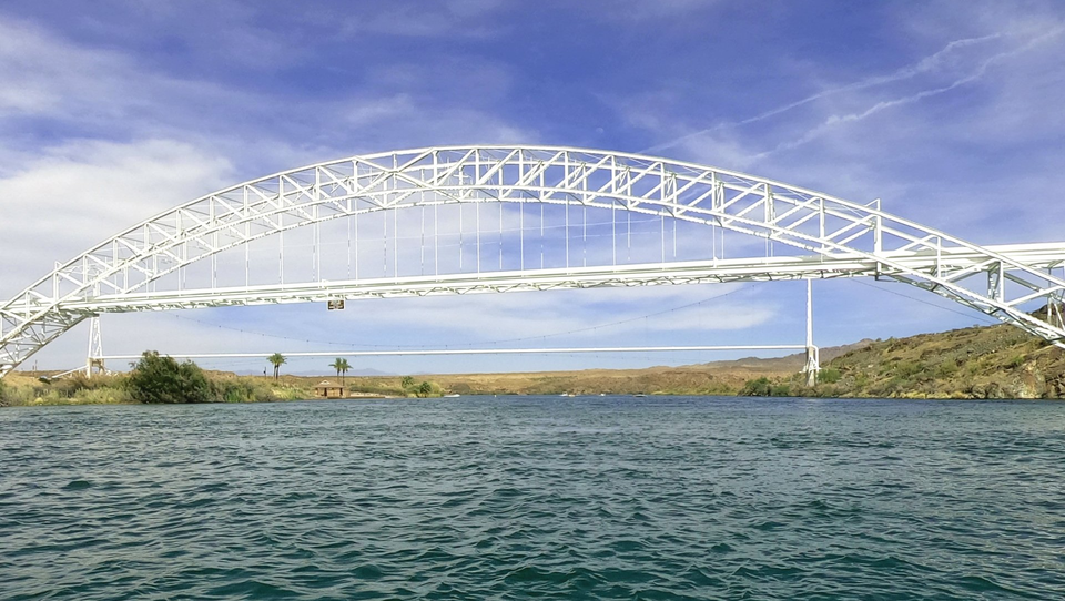 A white, arched steel bridge spans a wide river under a bright blue sky with wispy clouds.