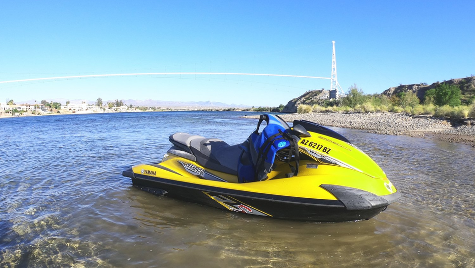 A yellow and black jet ski rests in the shallow, clear water of a river near a rocky shoreline on a sunny day.