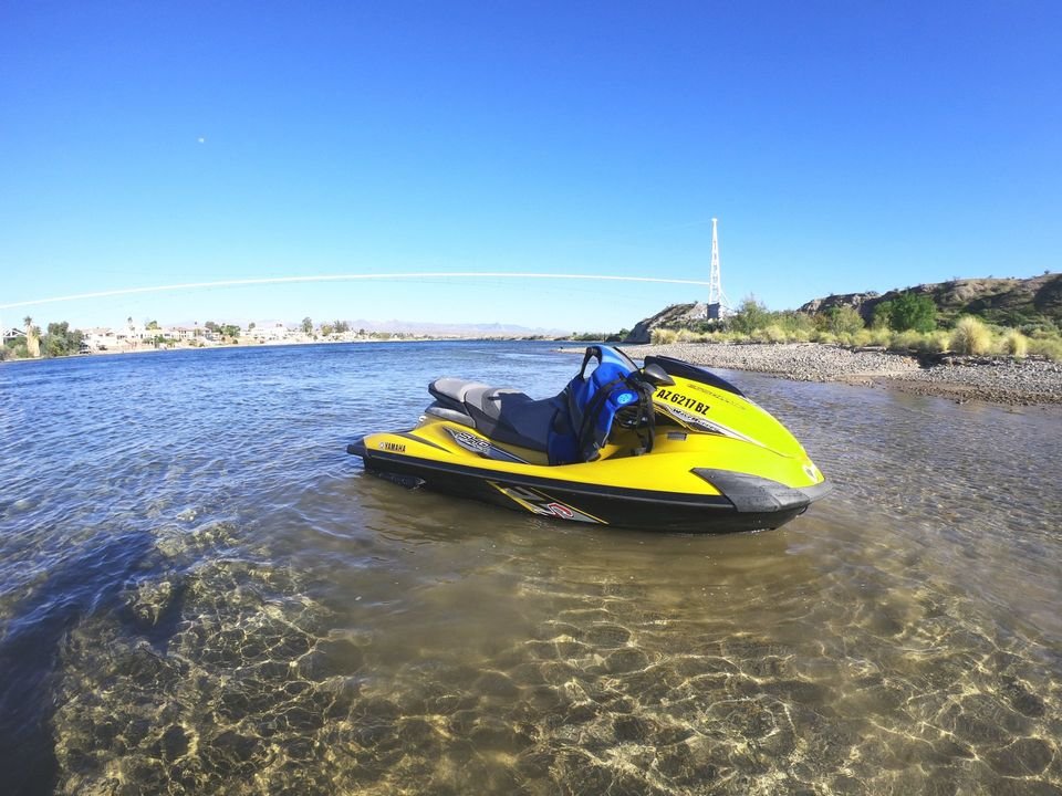 A bright yellow jet ski rests in shallow, clear water near a rocky shoreline under a bright blue sky.